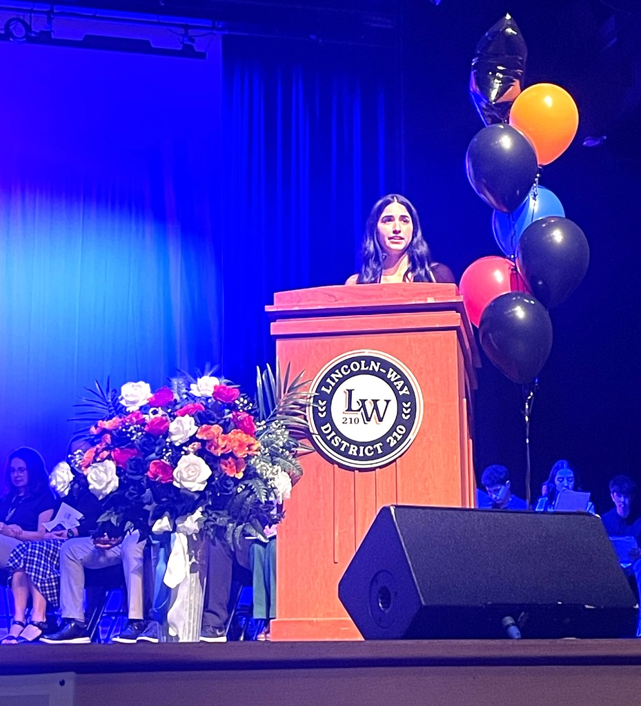 A student speaks at a podium with flowers, balloons, and a sign reading "Lincoln Way." Audience members are seated on a stage.