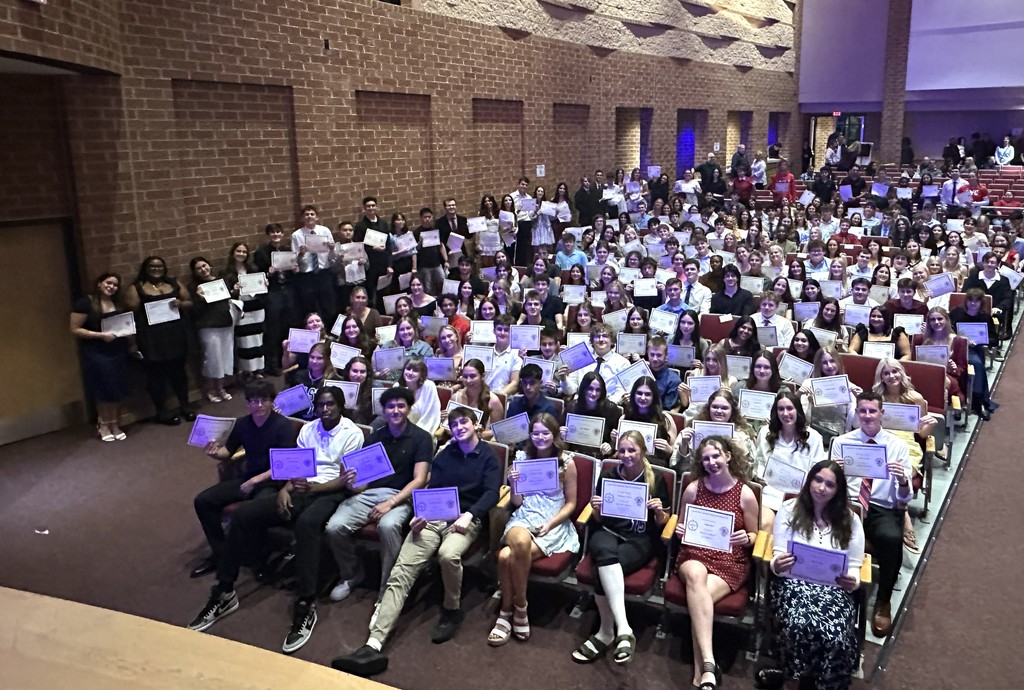 Large group of people holding certificates, seated in rows on a stage, brick wall in the background.