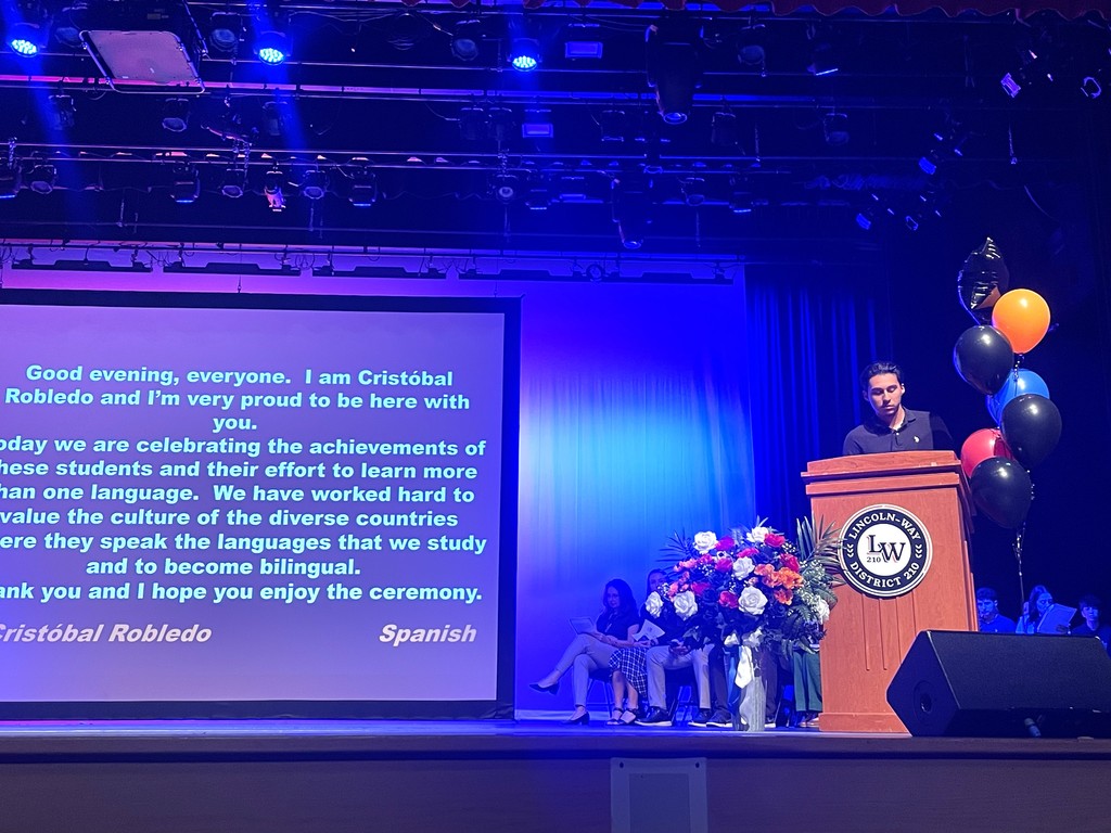 A stage with a person speaking, a screen with text, a flower arrangement, balloons, and a speaker.