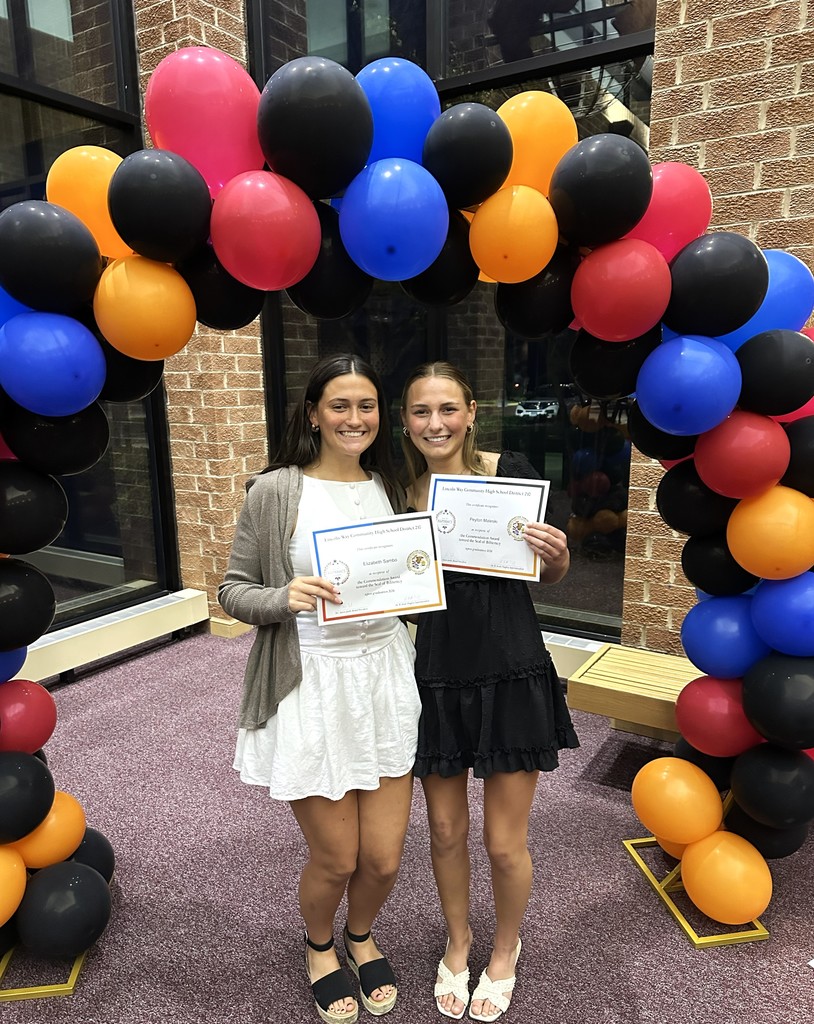 Two students  hold certificates before an arch of colorful balloons, with brick walls in the background.