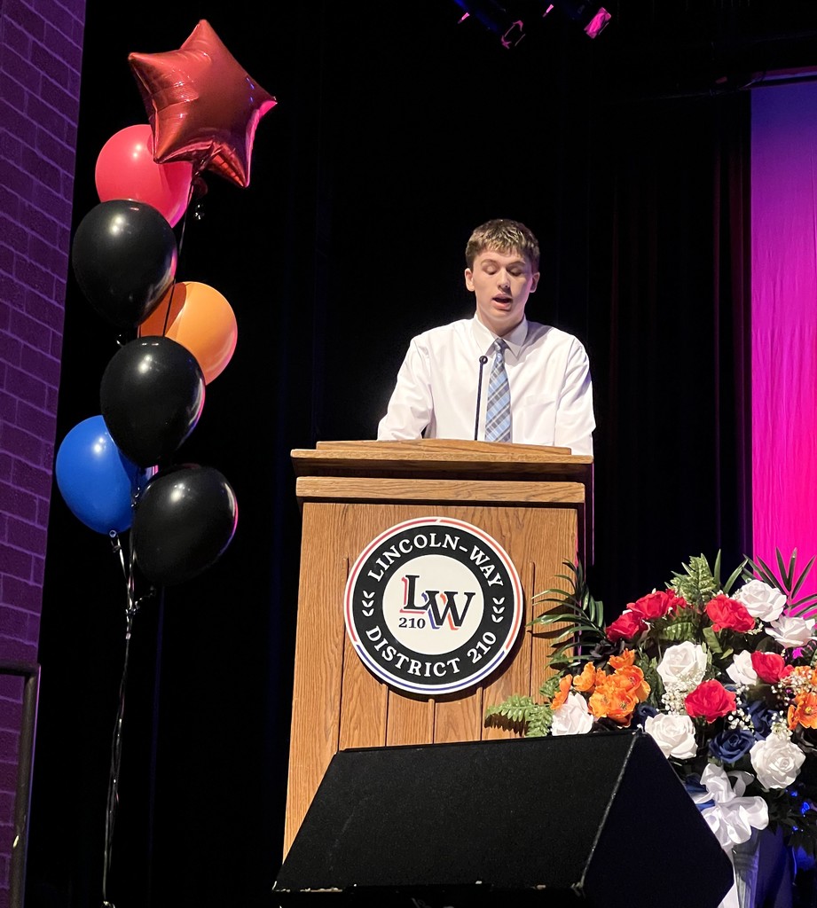 A podium with flowers and balloons stands in front of a speaker wearing a white shirt and tie.