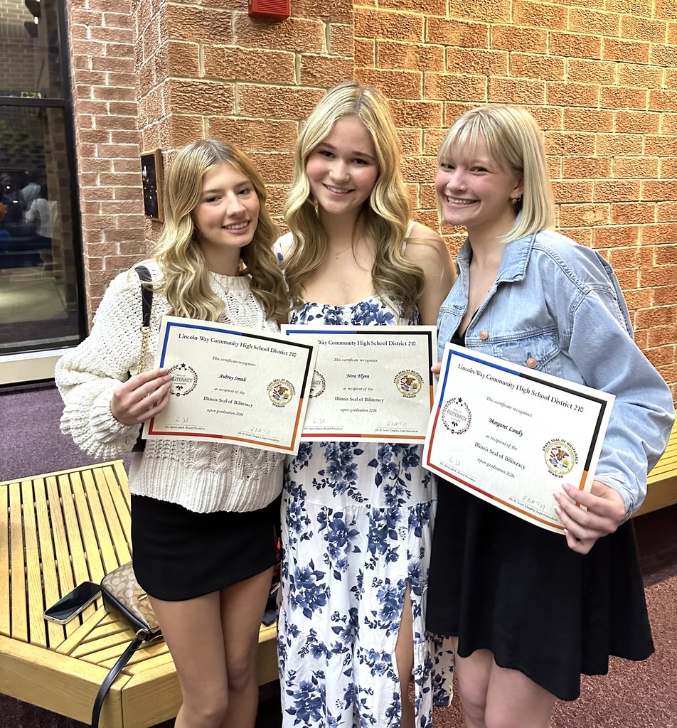 Three students  in a hallway pose for a photo, holding certificates. One wears a white sweater, another a floral dress, and the third a denim jacket.