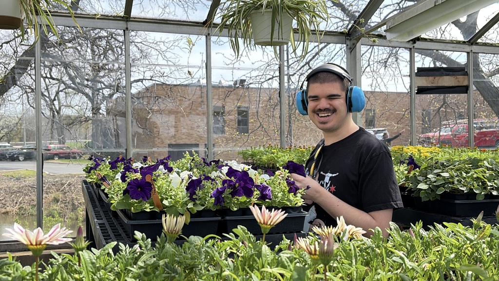 A person wearing headphones and a black t-shirt stands in a greenhouse with plants and flowers.