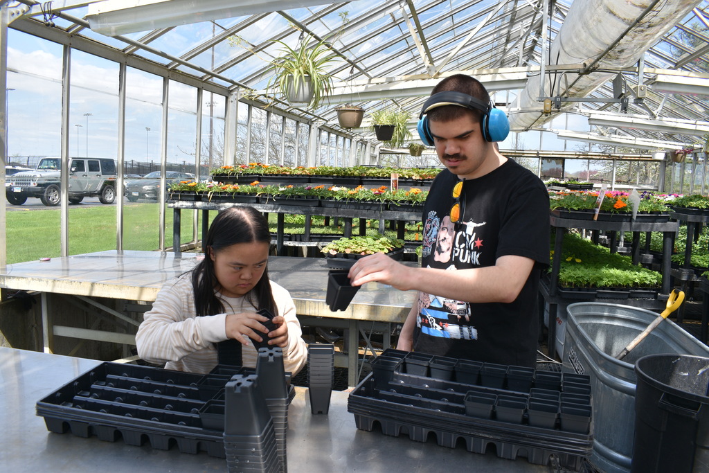 Two people in a greenhouse work with seedlings in pots; one wears headphones and the other wears a white long-sleeve shirt.