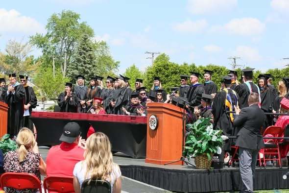 A graduation ceremony with graduates in caps and gowns on a stage, with faculty seated and a podium.