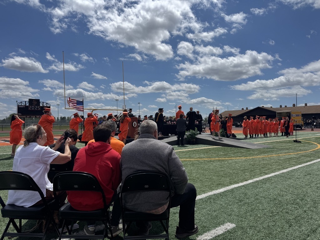 People seated on a football field, watching a group of individuals in orange uniforms on a stage.