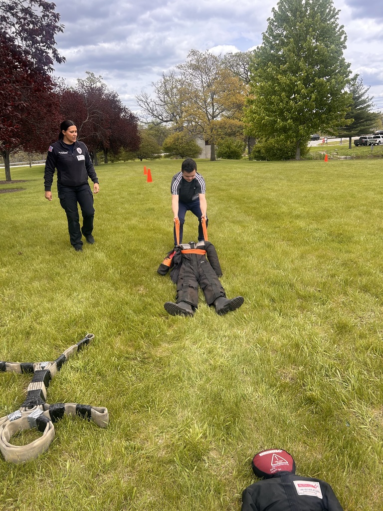 Two people are on a grassy field with trees. One person is pulling a dummy across the grass.
