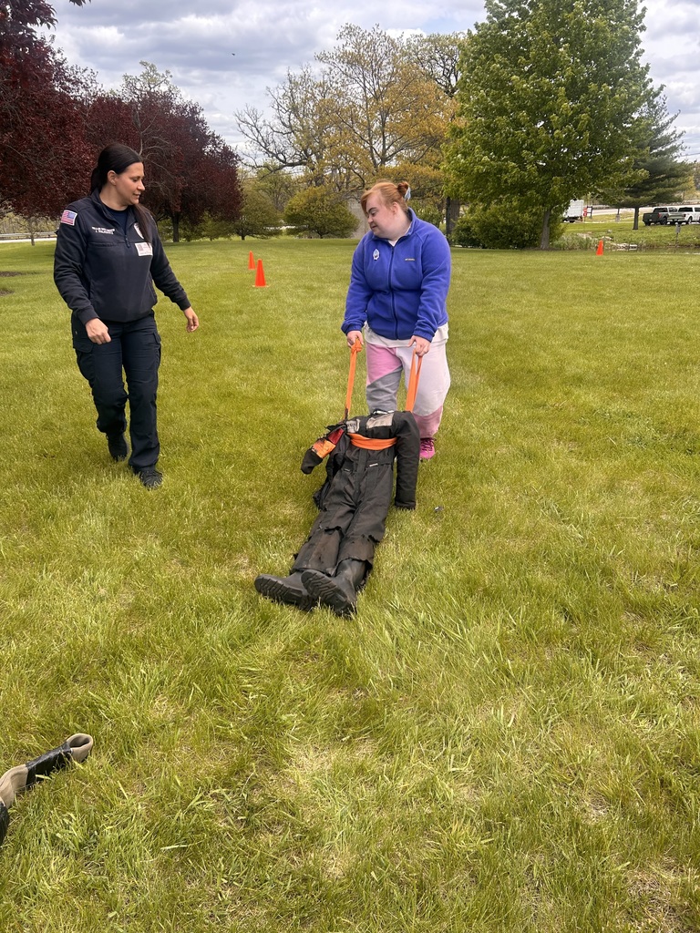 A student attempts a fire rescue on a dummy  in a black suit on a grassy field with trees and traffic cones.