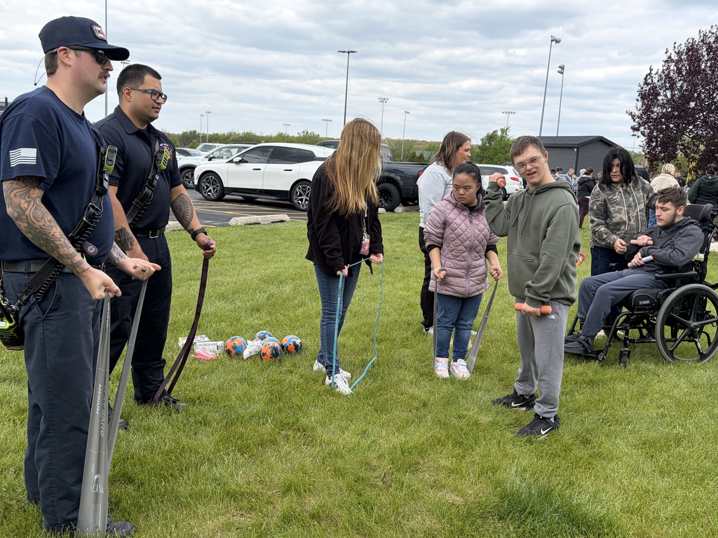 Several people in a grassy area, two firefighters are demonstrating an exercise with resistance bands to students.