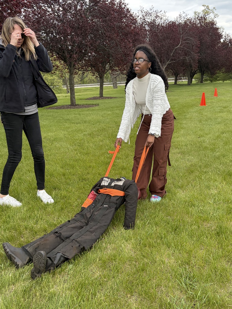 A student attempts a fire rescue on a dummy  in a black suit on a grassy field with trees and traffic cones.