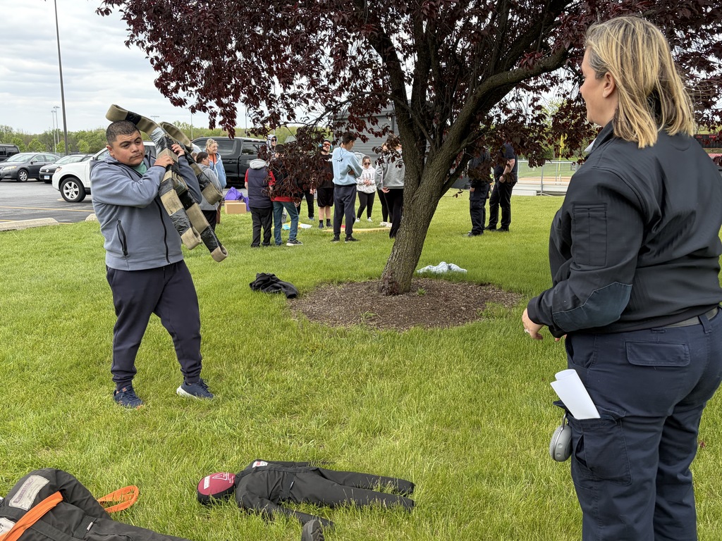A person in gray stands holding equipment; another person in black stands nearby. Both are outside with others.