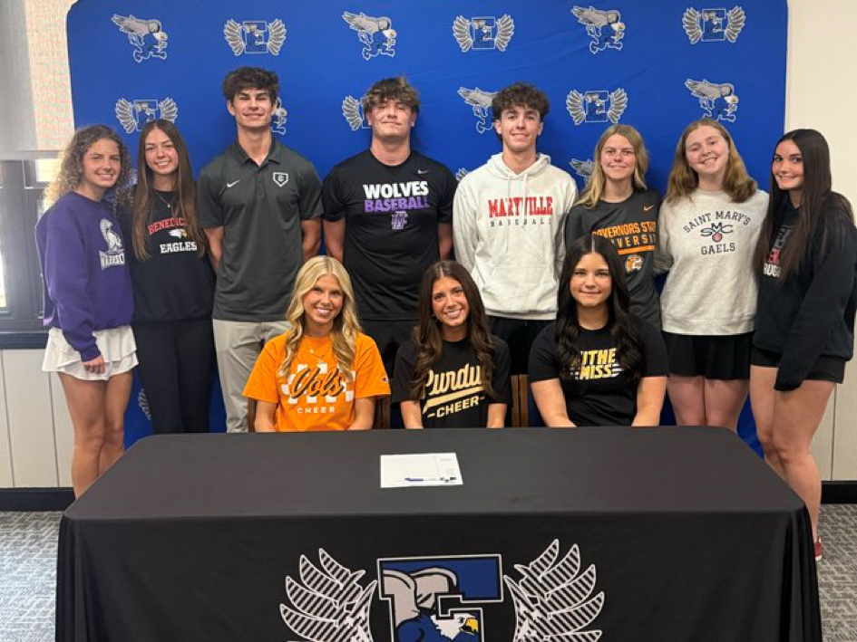 Group of young athletes and coaches standing behind a table, signing documents. A blue wall with logos is in the background.