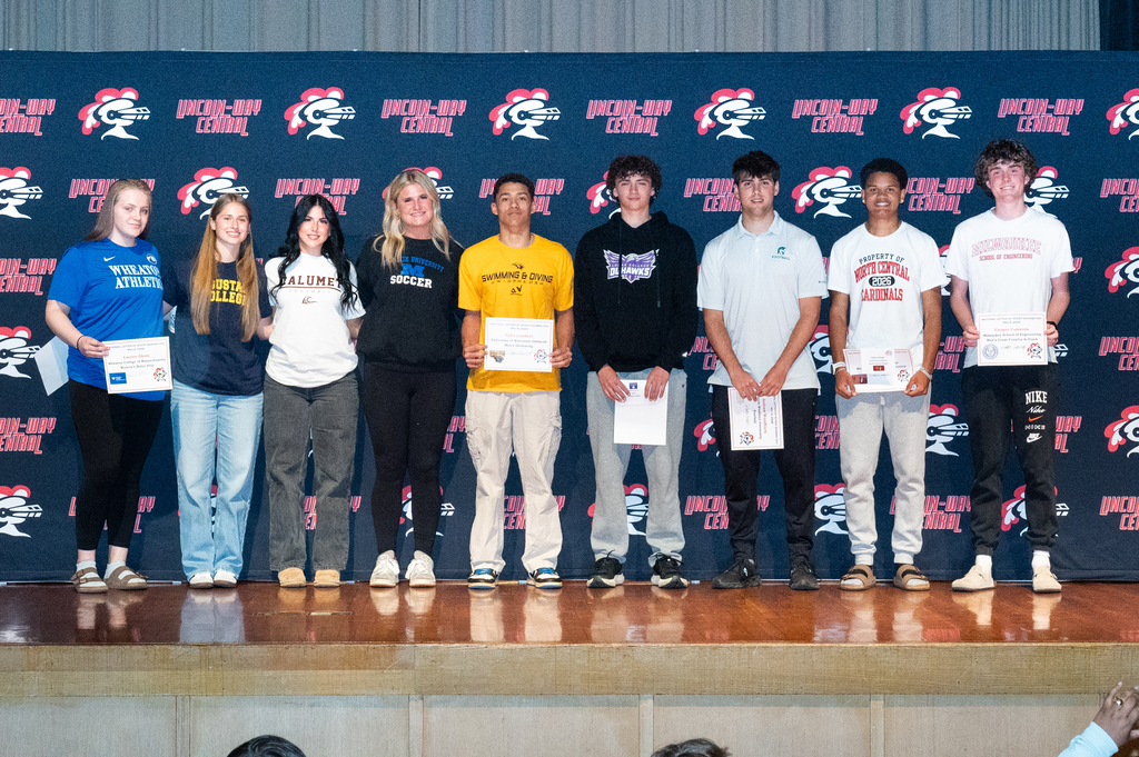 Nine individuals stand on a stage holding certificates; some wear sneakers, others boots. A blue backdrop displays white and red logos.
