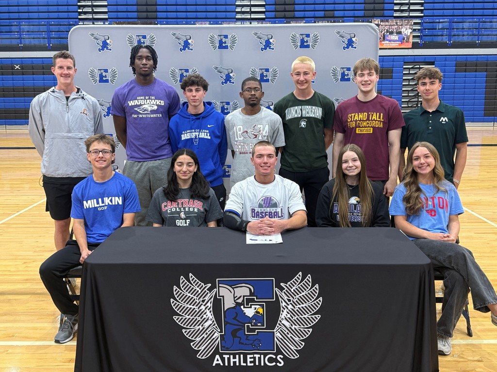 A group of young athletes and a coach posing in a gym with a table in front.