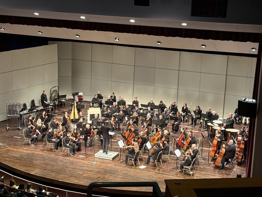 A large orchestra performs on a stage with wooden flooring, seated musicians, and a conductor. The stage is illuminated by overhead lights.