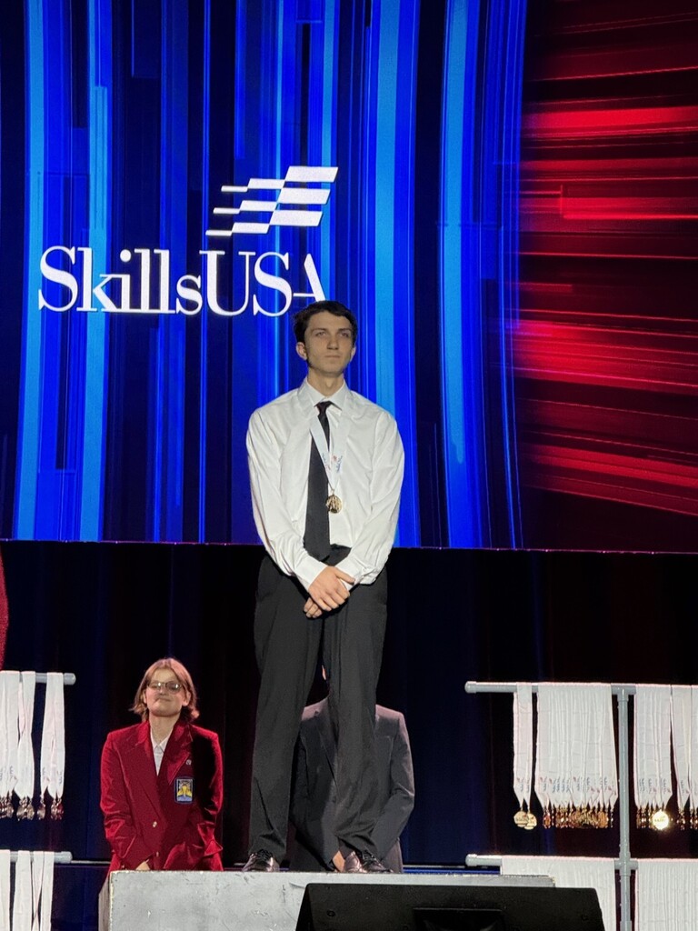Student stands on stage with two people behind him. Behind him is a backdrop with a SkillsUSA logo.