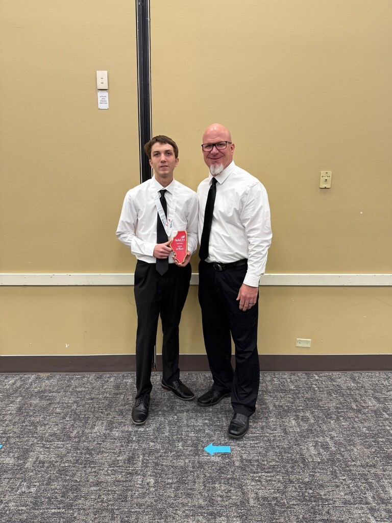 Two people in formal attire stand in a room with a beige wall. The student on the left holds a red award.