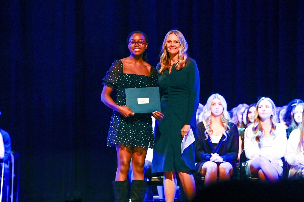 Two people stand on a stage holding a certificate, with a group of people sitting in the background.