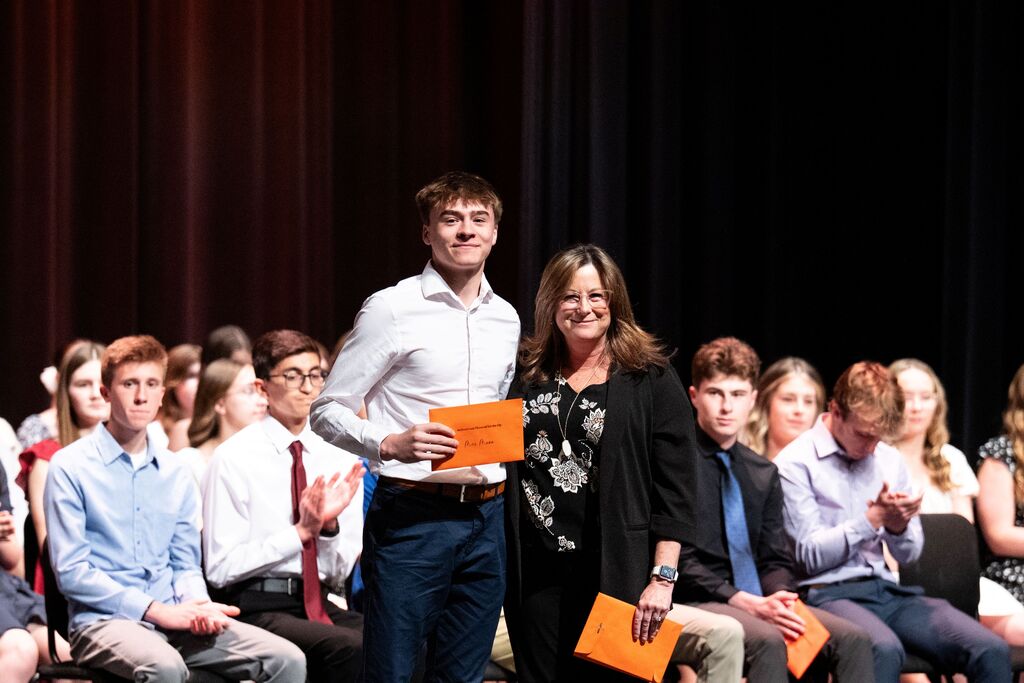 A group of students watch a young man receiving an award from a woman during a ceremony.