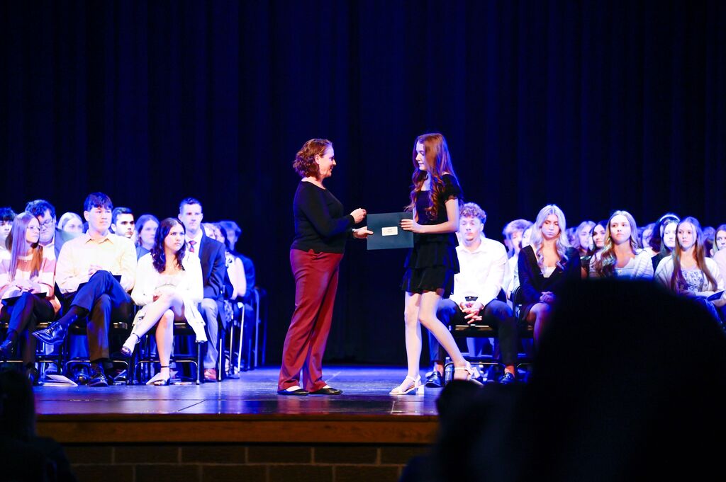 A person hands an envelope to another person on a stage. An audience sits in the background.