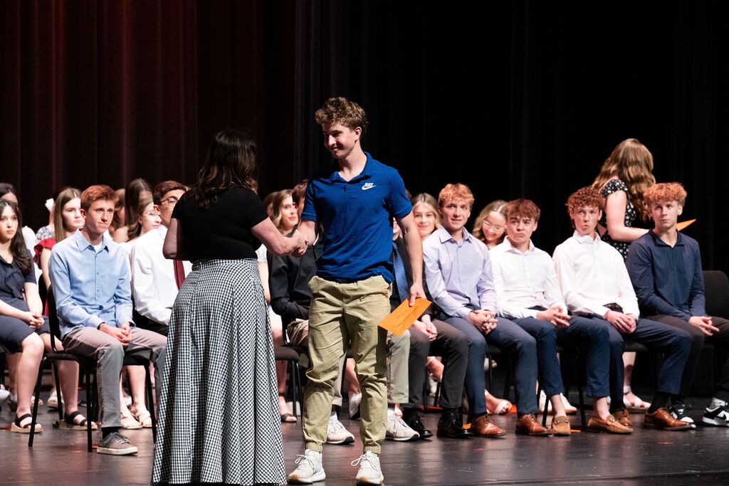 A person hands a certificate to another on a stage while an audience of seated people watch.