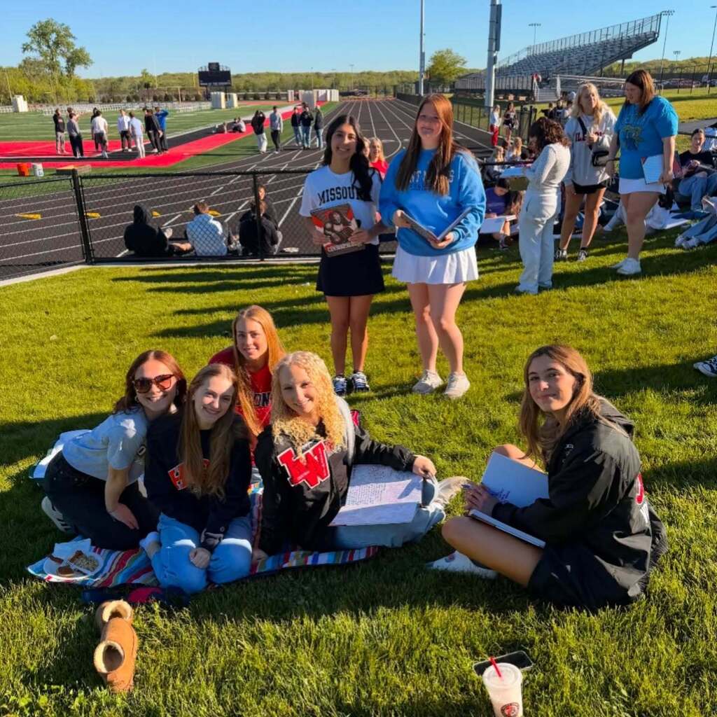 Group of girls sitting on a blanket on a grassy field, with a track and stadium in the background.