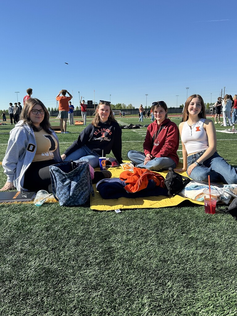Four students seated on a blanket on a field with various items around them. A clear blue sky in the background.