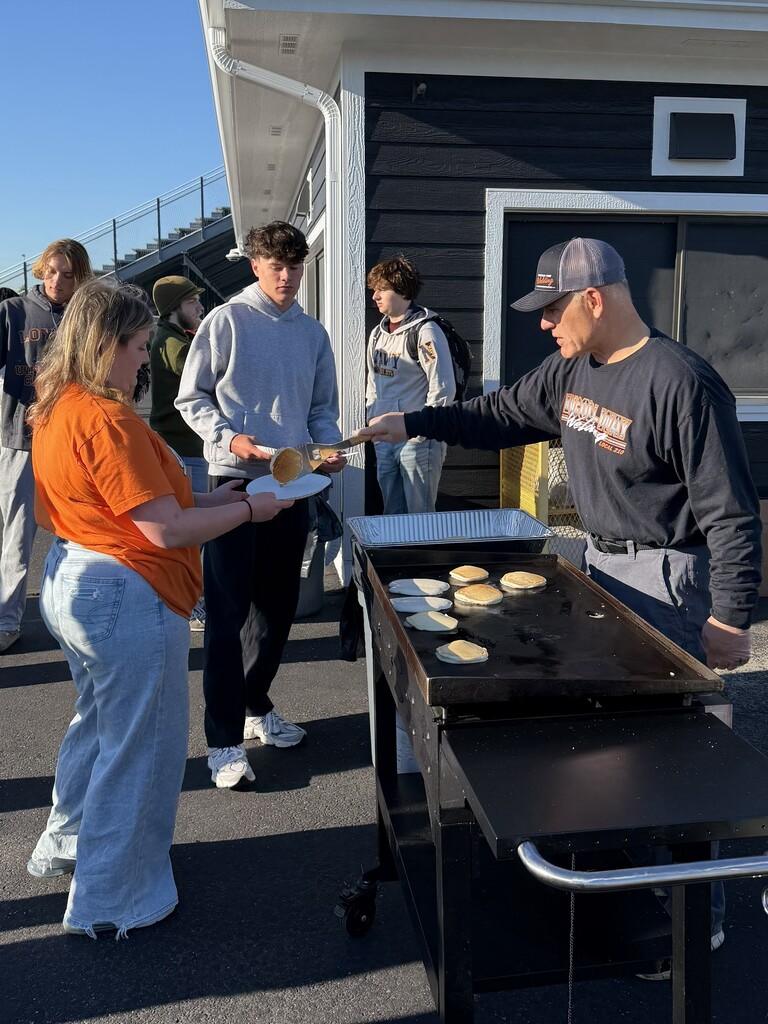 Two people in front of a building, one hands out pancakes on a grill to others.