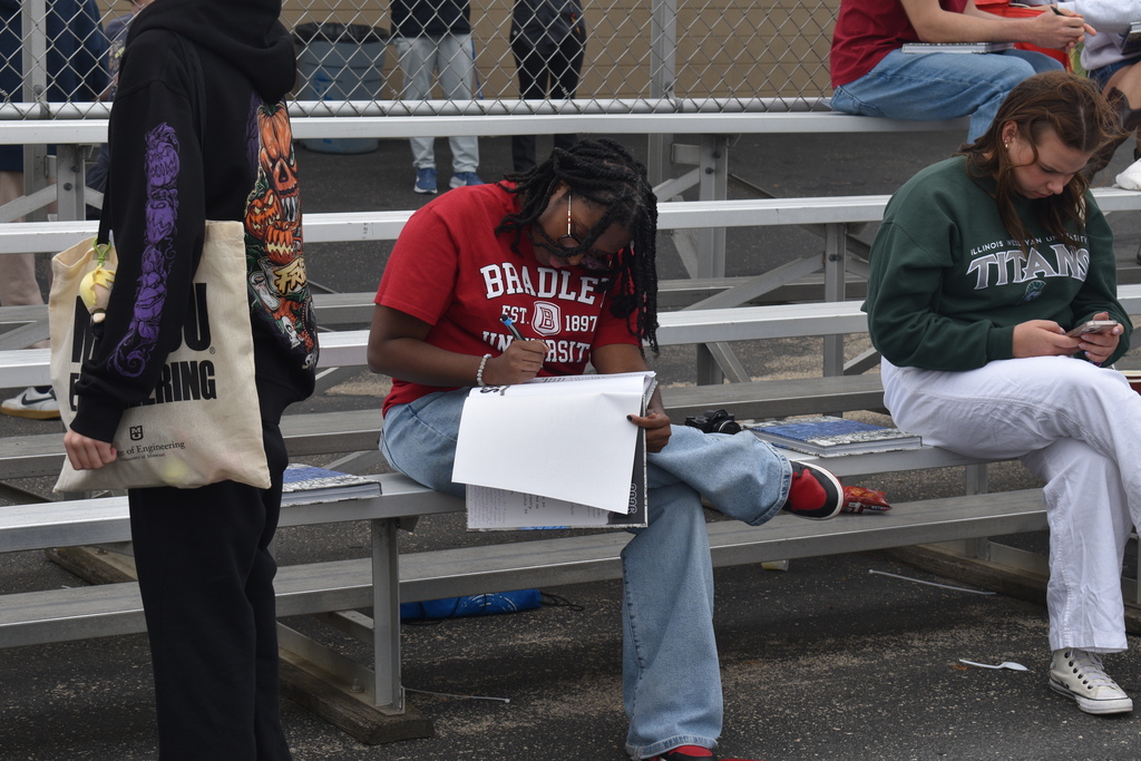 Two students seated on stadium bleachers, one writing on paper, the other using a phone.