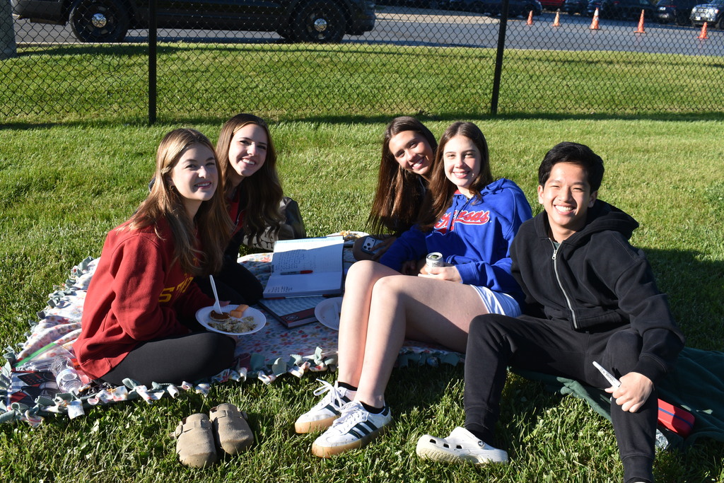 Six individuals seated on a blanket on a grassy field, smiling. One person holds a plate.
