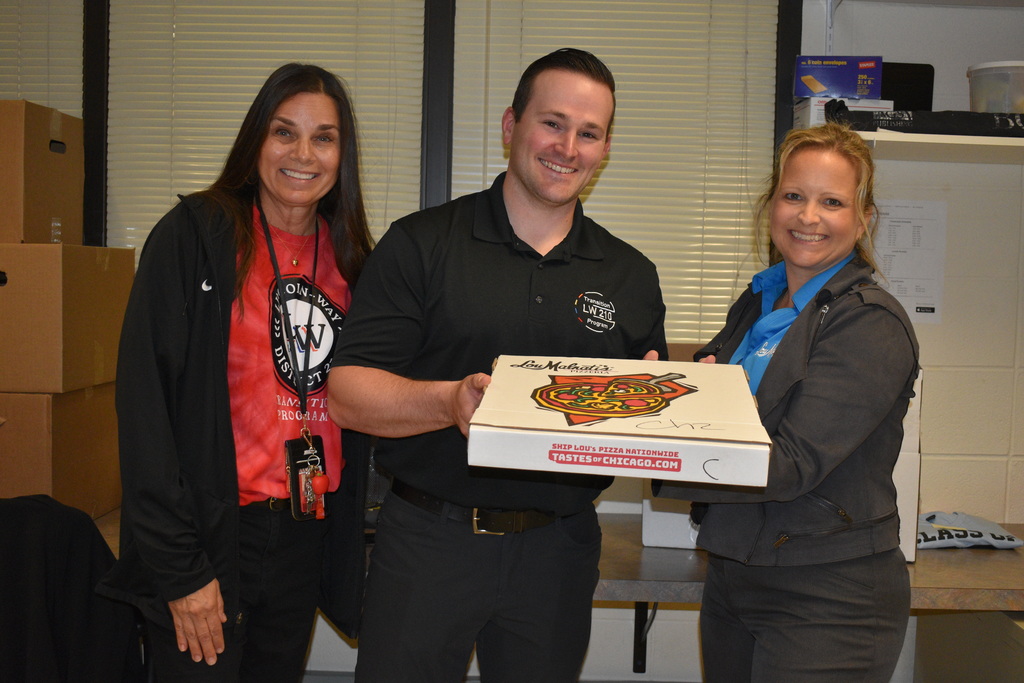 Three adults stand in a room, holding a pizza box. Two women in jackets, one in red and one in blue, flank a man in black.