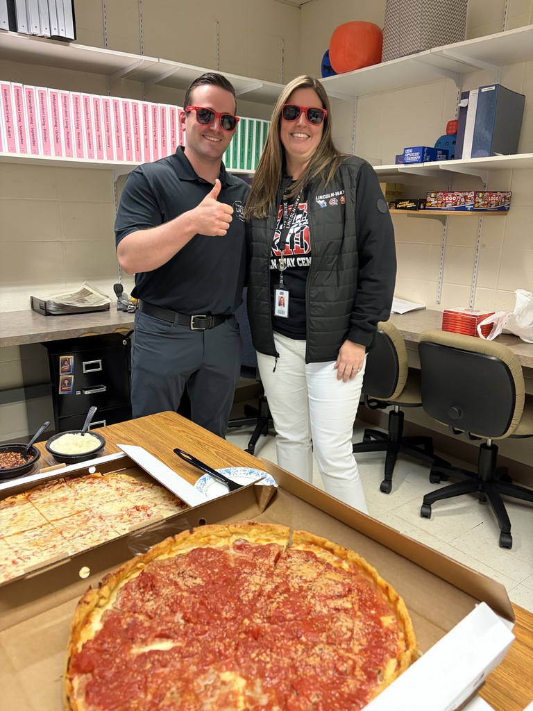 A man and woman stand behind a large pizza box in a room with chairs and shelves.