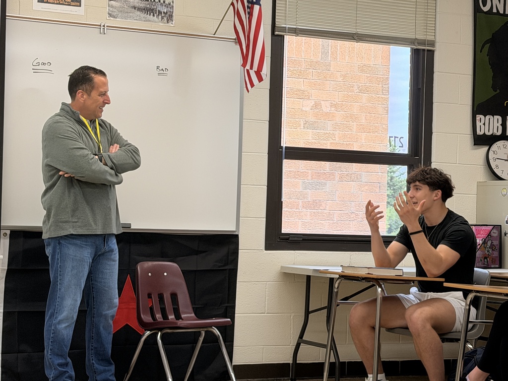 A man and a student in a classroom engage in conversation, with the student seated at a desk.