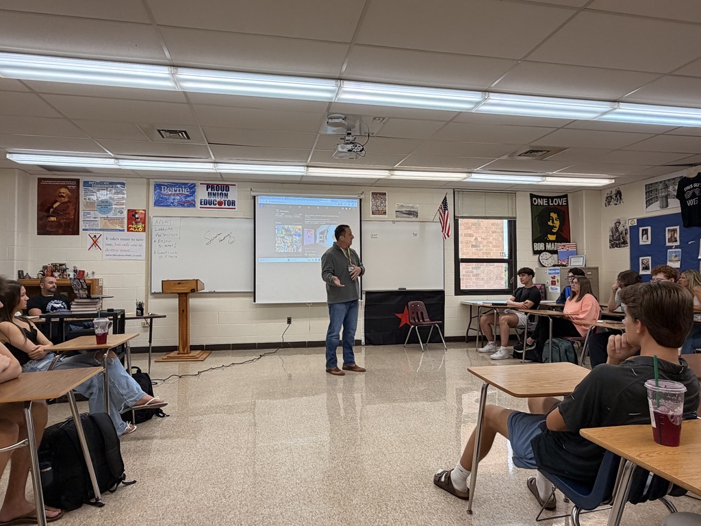 A classroom with white walls. Senator Hastings stands before a projector screen. Students sit at tables.