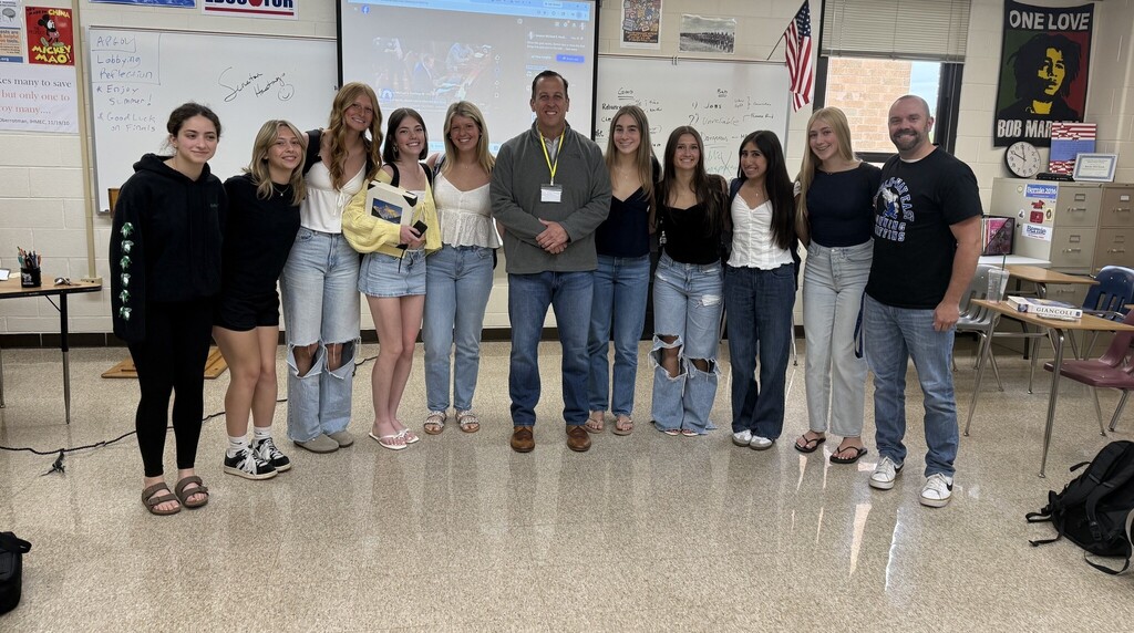 A group of students and two adults stand in a classroom, smiling, with whiteboards and a flag in the background.