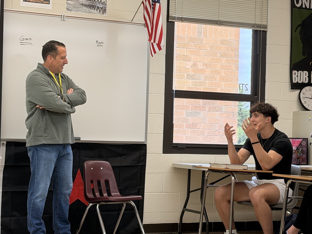 A teacher stands in front of a student seated at a table in a classroom. A clock and American flag are visible.