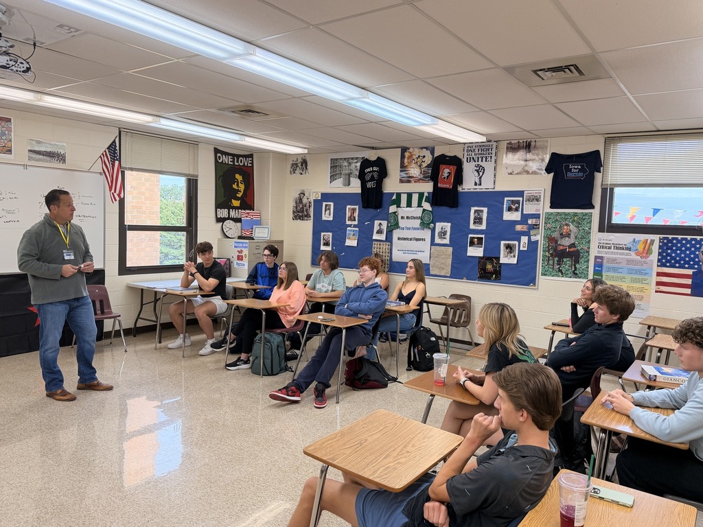 Sen. Hastings stands in front of class with students seated at desks, engaged in discussion. Walls are decorated with posters and flags.