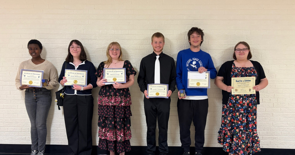 Six students stand together in front of a white wall, each holding a certificate. They wear casual clothing.