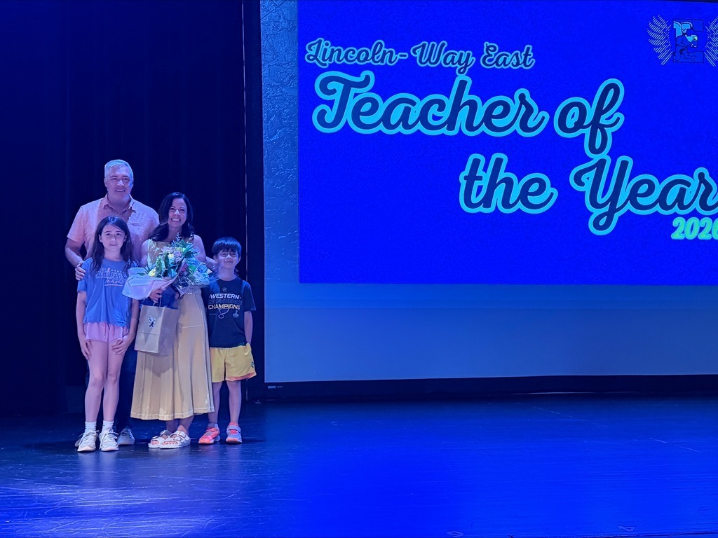 Five individuals stand on stage in front of a large screen displaying "Teacher of the Year 2022" in blue.
