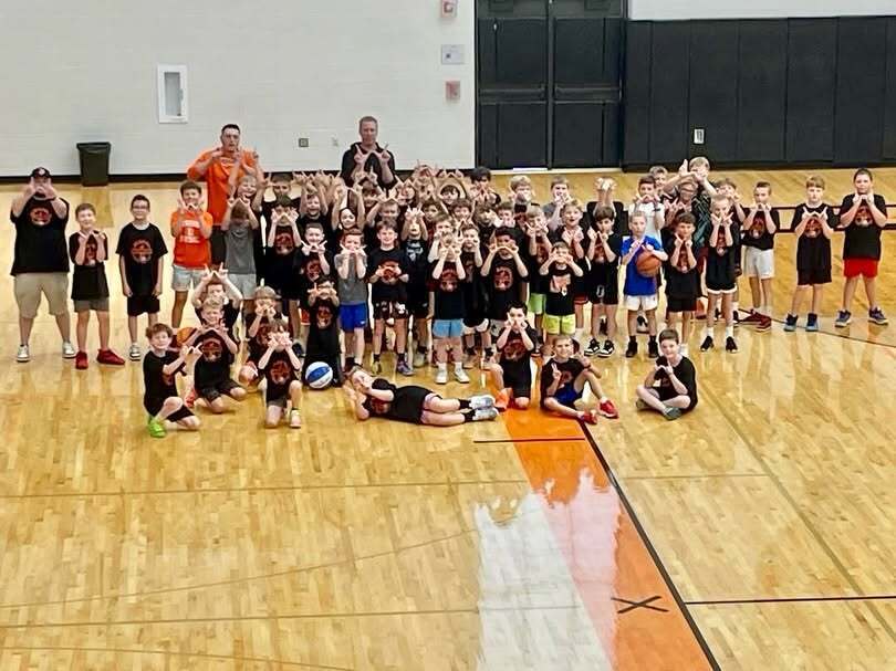 A large group of children and adults pose for a photo on an indoor basketball court, some sitting or standing.