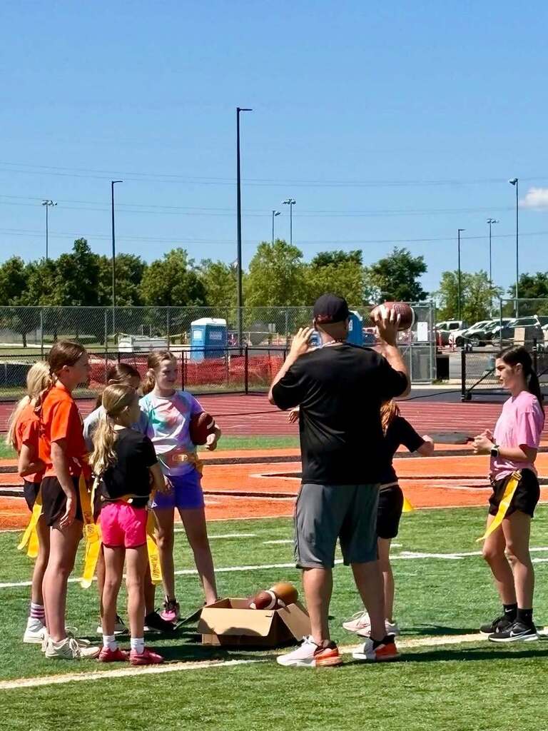 Nine children in different colored t-shirts and shorts, with yellow flags, gather around a coach on a football field.