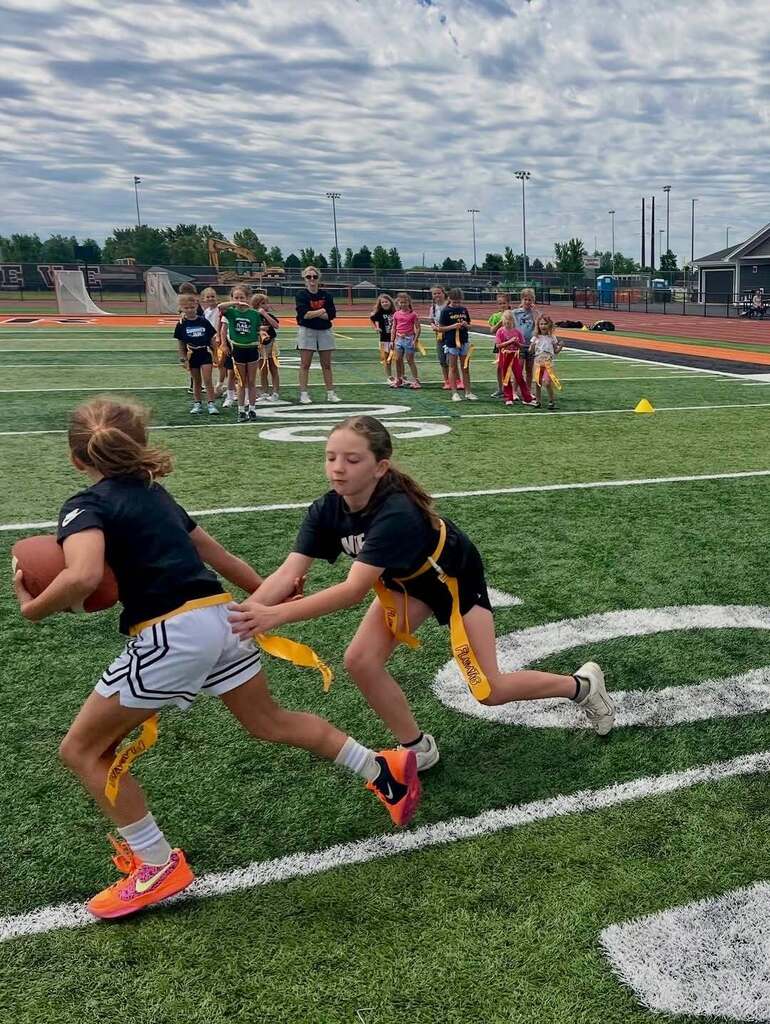 Two girls wearing black and white uniforms play flag football on a field. Other players and spectators are in the background.