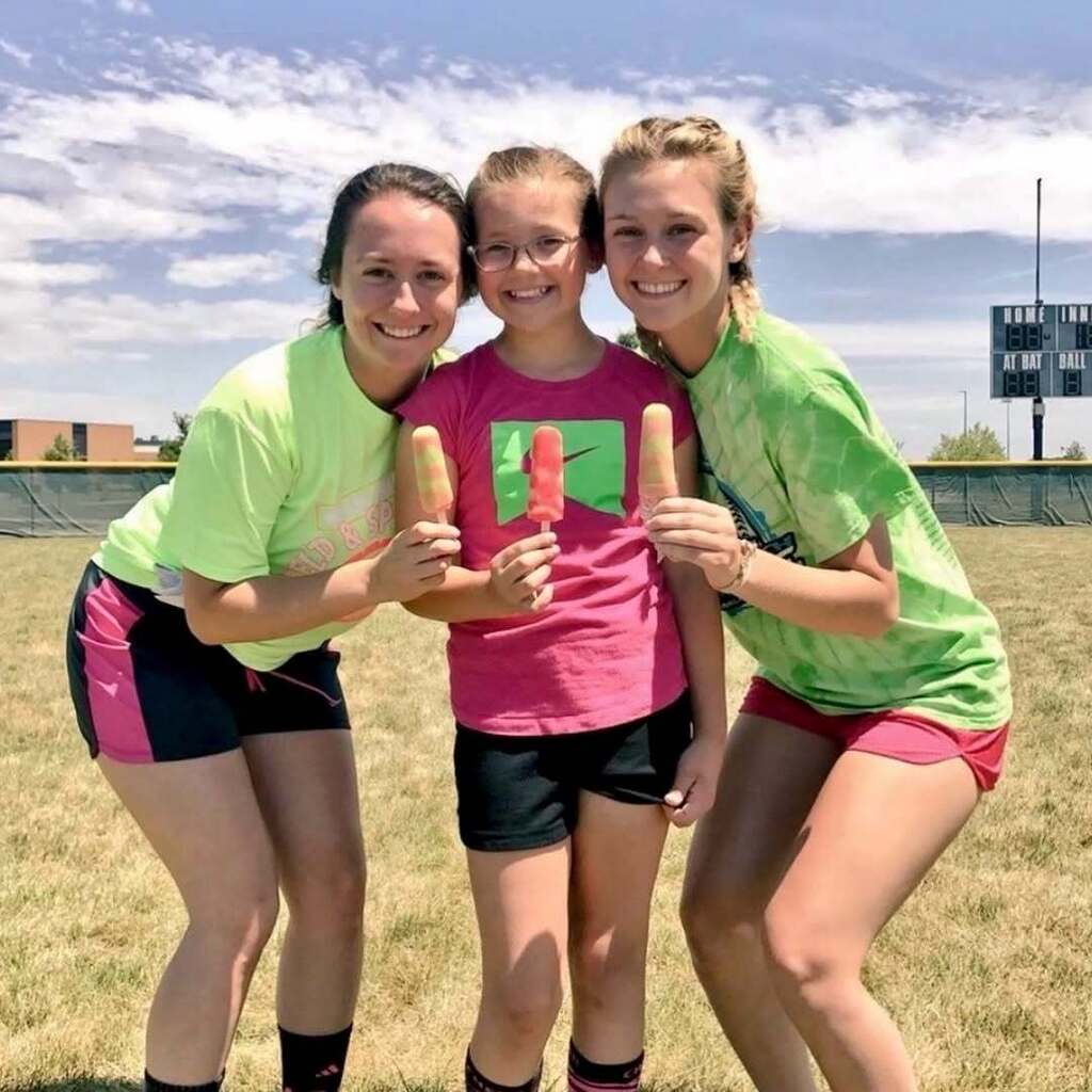 Three girls stand in a grassy field. They hold popsicles. They wear matching shirts and shorts.