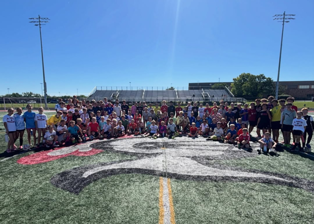 Many people gather on a football field with a white and black logo. A stadium is in the background.