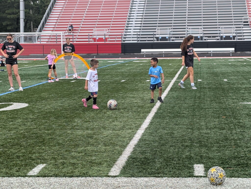 A group of young children play soccer on a field. Two kids are kicking the ball.