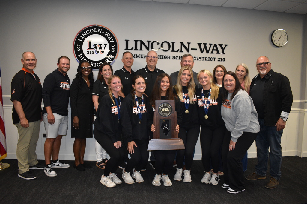 Group of adults and teens in matching black athletic wear, posing with a large trophy. Background shows a sign.