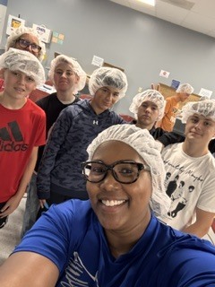 A group of people wearing white hairnets are smiling for a selfie. The background is a room with a gray wall and bulletin board.