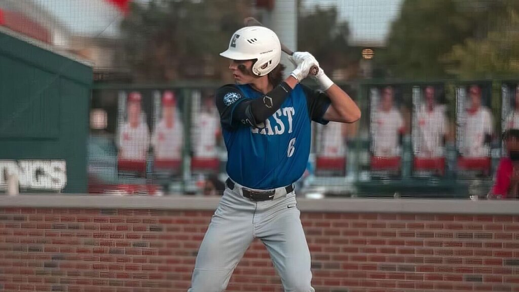 Baseball player in blue jersey with white helmet swings bat in a stadium. Brick wall with "NGS" behind player.