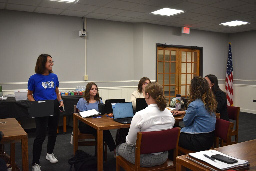 Several teachers seated around a desk, one woman stands at a table with a laptop, an American flag hangs behind them.
