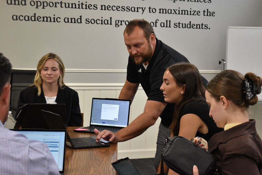 People are gathered around a table with laptops. A man is looking at the screen of the laptop.